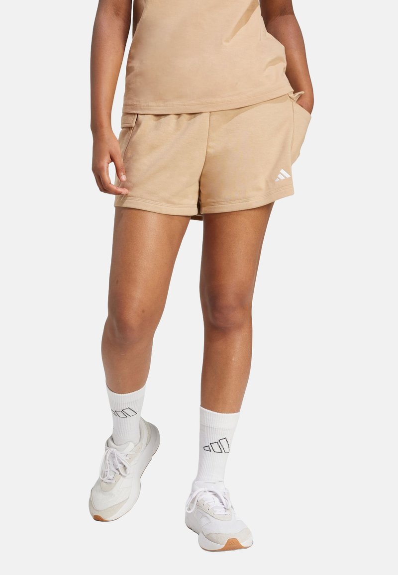 Beige sportovní šortky s elastickým pasem a postranními kapsami, v kombinaci s bílými kotníkovými ponožkami a světlými teniskami.