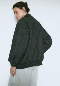 Mørkegrå herringbone bomberjakke med ribbede kanter og manchetter, der har en kontrasterende mørk krave og sidelommer.