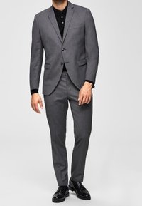Gray suit made of textured fabric with a tailored fit, paired with a black collared shirt. Accented with a single chest pocket and sleek black shoes.