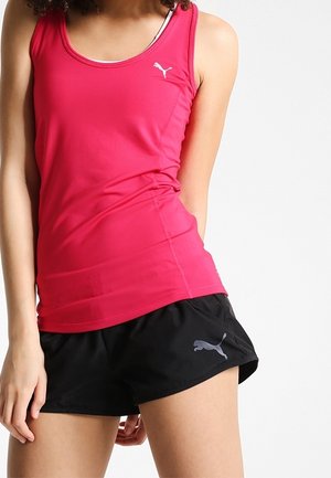 Vrouw die een nauwsluitend roze mouwloos Puma-shirt draagt en losse zwarte Puma-shorts, staand tegen een eenvoudige lichte achtergrond.