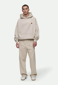 Beige ykkösluokan huppari, jossa on pieni punainen logo, yhdistettynä vastaaviin beigeihin collegehousuihin. Molemmat on valmistettu pehmeästä kankaasta. Valkoiset kengät viimeistelevät asun.