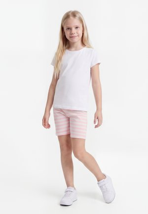 Νεαρό κορίτσι στέκεται, φορώντας λευκό μπλουζάκι, ροζ-άσπρα ριγέ σορτς και λευκά αθλητικά παπούτσια πάνω σε ένα απλό φωτεινό φόντο.