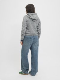 Un sweat à capuche gris en tissu doux, associé à un jean en denim bleu ample. L'arrière-plan est blanc uni, mettant en valeur la tenue.