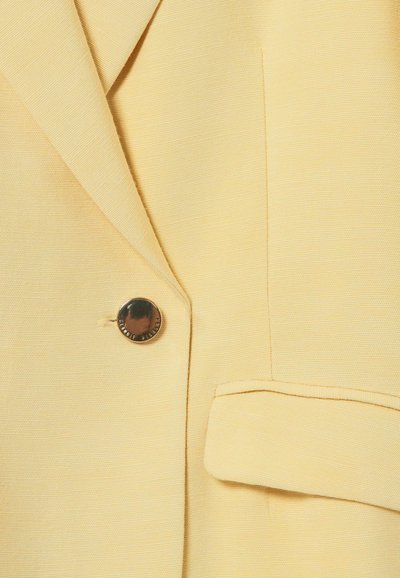 Blazer giallo chiaro in tessuto texturizzato, con un singolo bottone marrone, rever affilati e una tasca sul davanti.
