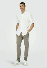 Witte overhemd met korte mouwen, grijze op maat gemaakte broek en witte sneakers met accenten. Model staat met handen in de zakken tegen een lichte achtergrond.