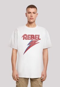 Baltas kokvilnas T-krekls ar lielu, novecojušu sarkanu "REBEL" tekstu un zilu zibens bultu grafiku. Brīva piegriezne un īsas piedurknes.