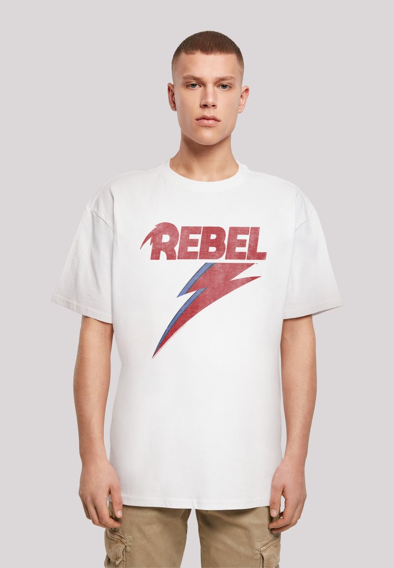 Baltas kokvilnas T-krekls ar lielu, novecojušu sarkanu "REBEL" tekstu un zilu zibens bultu grafiku. Brīva piegriezne un īsas piedurknes.
