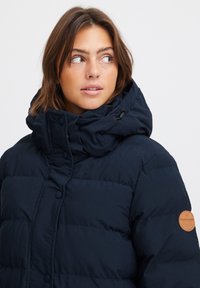 Marineblå dunjakke med høy krage, knappelukking, og en lærlogo på venstre erme. Glatt tekstur, isolert design.