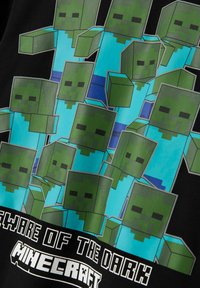 Melnā T-kreklā ir attēloti vairāki zaļi kvadrātiem līdzīgi zombiju tēli zilās krekls, apakšā ir teksts "Uzmanieties no Tumsas" un "Minecraft".