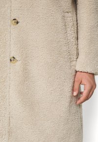 Cappotto di lana beige texturizzato con grandi bottoni e la mano destra di una persona appoggiata accanto alla tasca del cappotto.