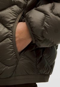 Ręka wsunięta w boczną kieszeń ciemnooliwkowej pikowanej kurtki puchowej, ukazująca teksturę i szczegóły szycia.