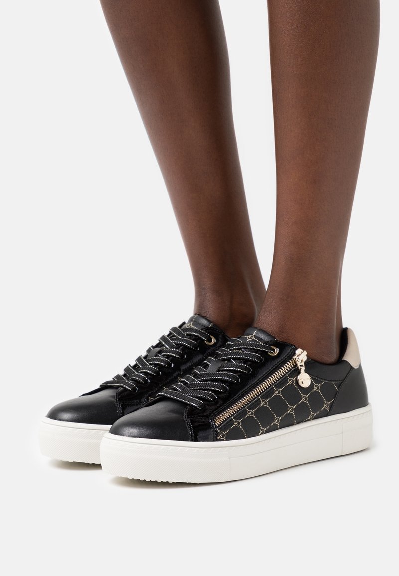 Baskets en cuir noir avec zip décoratif, semelle blanche et motif texturé sur les côtés avec des accents dorés. Les lacets sont sombres et ronds.
