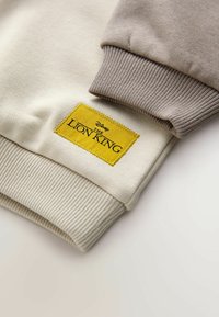 Helle beige und taupe Sweatshirtärmel mit gerippten Bündchen. Mit einem gelben gestickten Aufnäher mit der Aufschrift "Der König der Löwen." Glatte Textur.