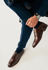 Brown leather dress shoes with brogue detailing. Worn with navy textured trousers rolled at the cuffs and navy socks.