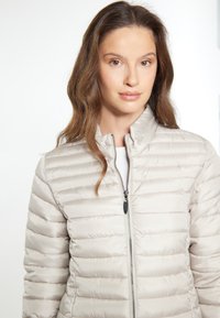Lichtbeige pufferjack met horizontale gestikte ontwerp, hoge kraag, voorrits en glanzende textuur. Geschikt voor casual draagmode.
