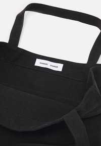 Black canvas tote bag with a rectangular shape, featuring two sturdy shoulder straps and a white brand label stitched on top.