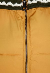 Gul vadderad jacka med en svart dragkedja i mitten, med en grön stickad krage med vita zigzag-mönster.