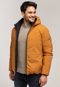 Pomarańczowa kurtka puchowa z czarnym wnętrzem i kapturem. Posiada zamek błyskawiczny z przodu oraz naszywkę z logo na rękawie. Nosi się ją na jasnym swetrze.