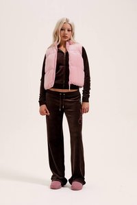 Ρόζ αμάνικο μπουφάν puffer πάνω από καφέ βελουτέ φόρμα. Η φόρμα έχει τοπ με φερμουάρ, παντελόνι με κορδόνι και κεντήματα. Ρόζ παπούτσια τύπου slip-on.