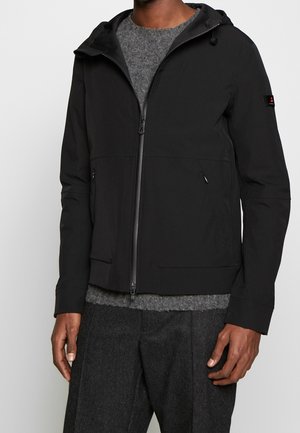 Homme portant une veste noire à capuche zippée par-dessus un pull gris avec un pantalon gris foncé sur un fond uni.