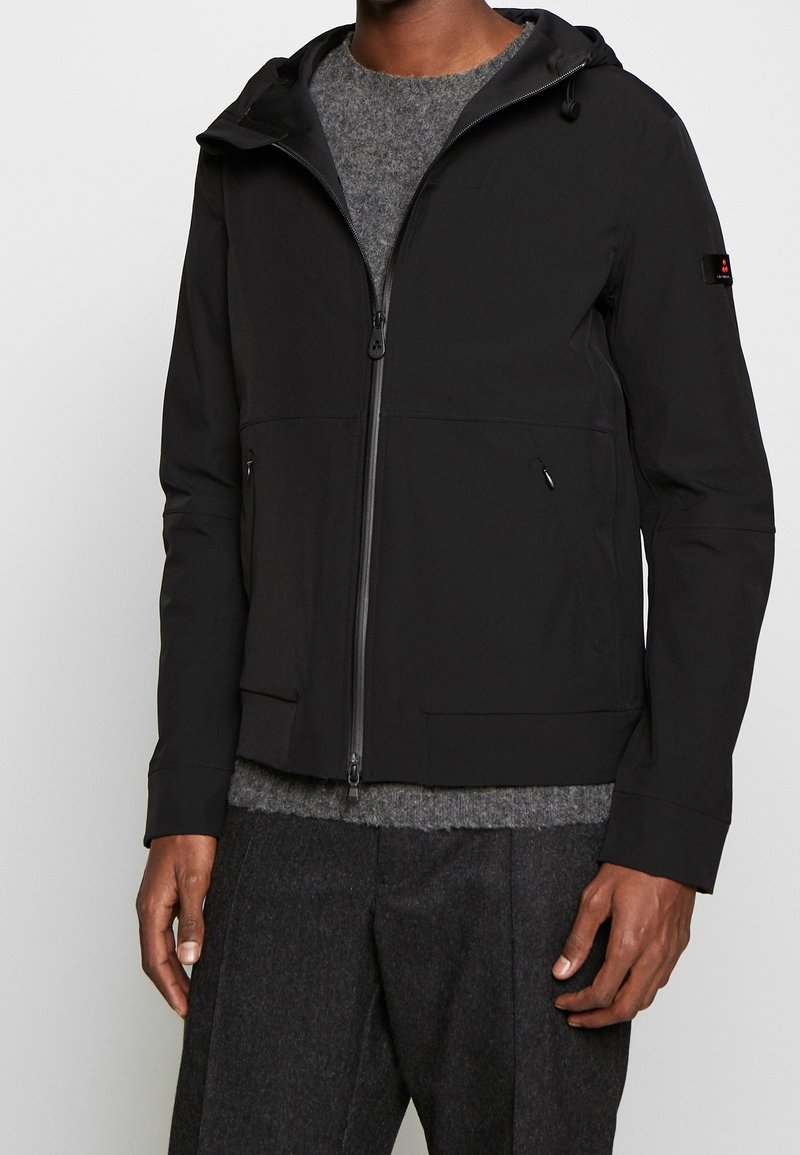 Homme portant une veste noire à capuche zippée par-dessus un pull gris avec un pantalon gris foncé sur un fond uni.