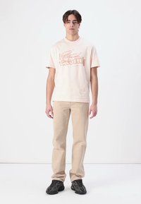 Camiseta de algodón rosa claro con un logo de cocodrilo y el texto "LACOSTE", combinada con pantalones beige y zapatillas negras, posando frente a un fondo blanco.
