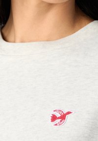 Szara bluza z okrągłym dekoltem i małym, czerwonym haftowanym logo ptaka na piersi, wykonana z miękkiej bawełny.