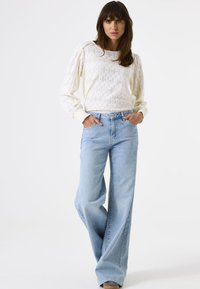 Mujer vestida con una blusa blanca de textura de manga larga y vaqueros anchos de un azul claro, de pie con las manos en los bolsillos contra un fondo blanco.