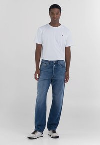 T-shirt blanche avec un petit logo rouge, associée à un jean en denim bleu ample et des baskets blanches avec des accents noirs. Fond uni.