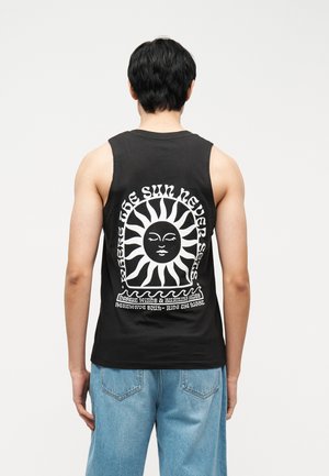Man met een zwart mouwloos shirt met een witte zonafbeelding en de tekst "The sun never sets" op de achterkant, gecombineerd met lichtblauwe jeans.