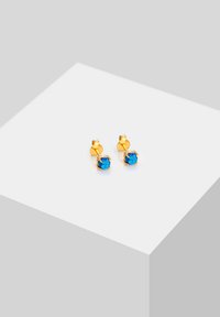 Boucles d'oreilles en métal doré avec des sertissages en cristal bleu en forme ronde, maintenues par un design de griffes simple. Dos en métal texturé visible.