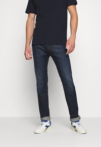 Granatowe dżinsy o slim fit z podwiniętymi nogawkami, w połączeniu z granatową koszulką i białymi sneakersami z niebieskimi akcentami.