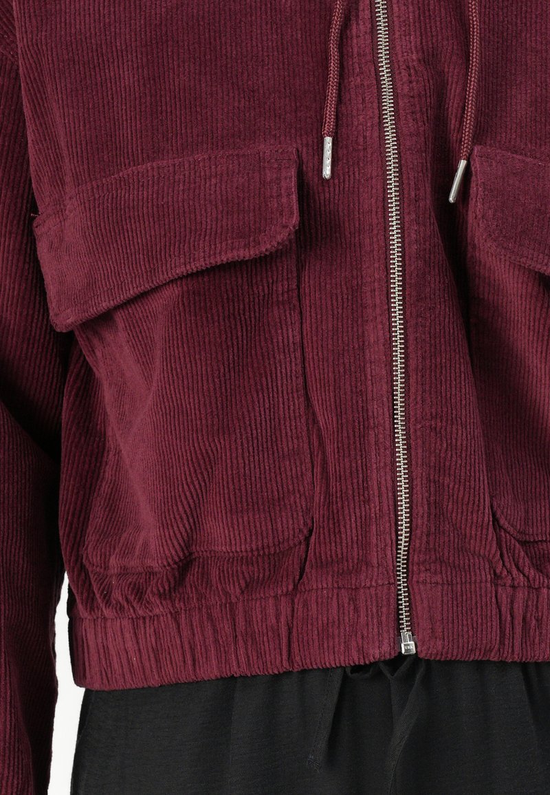 Bordowa kurtka z kordurowej tkaniny ma zamek z przodu, dwie naszywane kieszenie, ribbowe wykończenie oraz teksturowany materiał. Widoczna jest czarna tkanina poniżej.