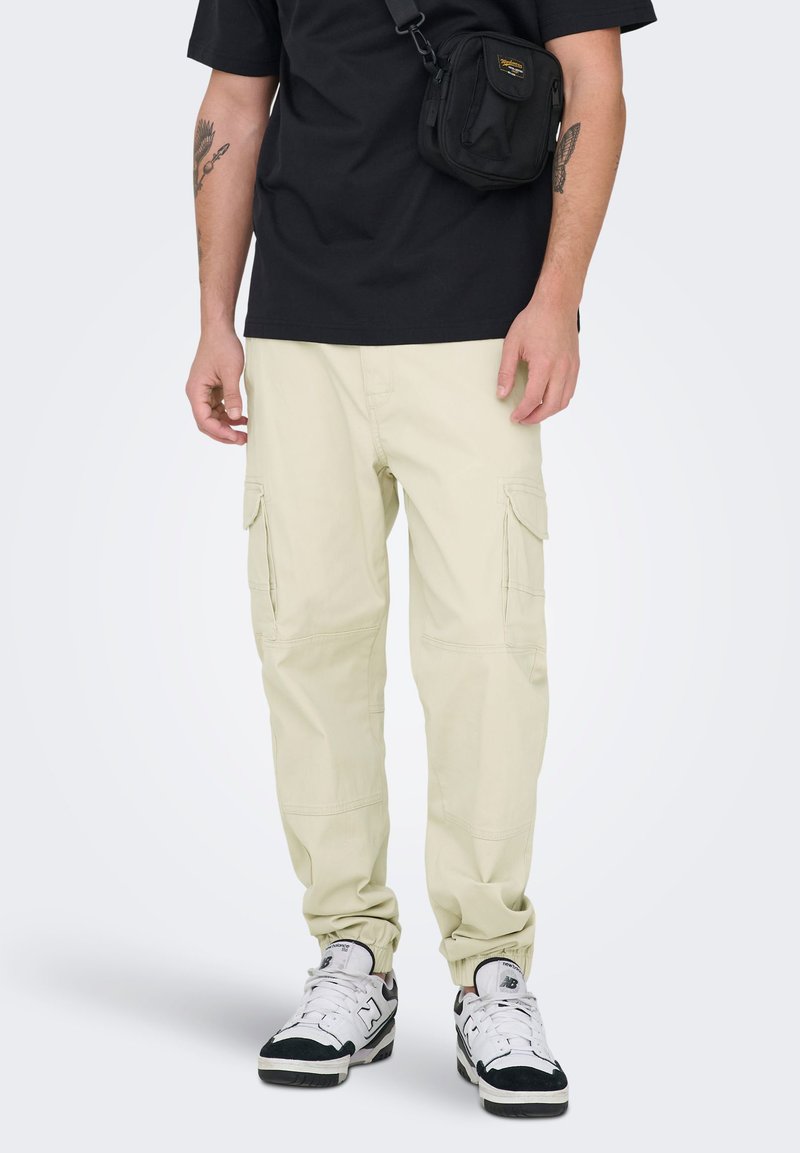 Pantalon cargo crème avec plusieurs poches latérales, poignets resserrés et coutures visibles, associé à un t-shirt noir et des baskets blanches.