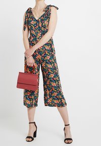 Jumpsuit floral avec un décolleté en V plongeant et des bretelles à nouer, présentant des fleurs multicolores sur un fond sombre. Associé à un sac à main rouge.