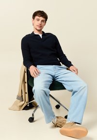 Tummansininen villapaita kauluksella, vaaleansiniset farkut ja vaaleat kengät. Malli istuu vihreällä tuolilla, jonka vieressä on beige takki.