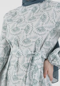 Kevyt mekko, jossa on kukkakuviointi vihreänä vaalealla taustalla, vyötäröllä on vyö ja pitkät puffatut hihat.