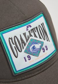 Szary czapka z kolorową haftowaną naszywką z napisem "KOALICJA", wzorem fali i rokiem "1993" poniżej. Naszywka ma turkusowe i fioletowe akcenty.