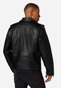 Černá kožená bunda s texturovaným povrchem, střih na míru a ramenními epolety, nošená přes tmavé džíny, zobrazena zezadu.