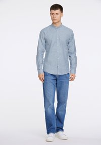 Camisa de botones azul claro con cuello mao, combinada con jeans azules y zapatillas blancas. La camisa presenta una textura sutil.