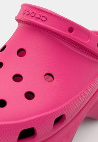 Rosa Crocs med en teksturert overflate, utstyrt med ventilasjonshull og en tykk, avrundet stropp for sikker bruk. Sålen har et mønstrete grep.
