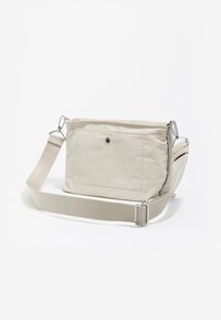 Beżowa torebka na ramię wykonana z miękkiego materiału, z zamkiem błyskawicznym na górze, regulowanym paskiem oraz przednią kieszenią z czarnym detalem guzika.