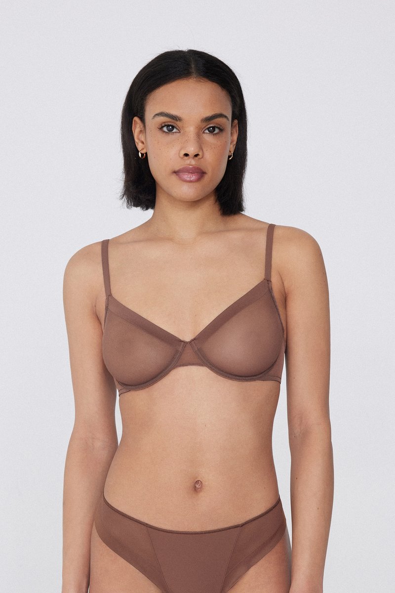Femme aux cheveux courts et foncés portant un soutien-gorge marron transparent assorti à sa culotte, debout devant un fond uni gris clair.