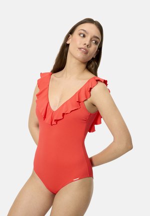 Mujer vistiendo un traje de baño sin mangas de color rojo brillante con escote en V y volantes en los hombros, posando con las manos detrás de la espalda sobre un fondo liso.