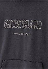 Sudadera oscura con "RHODE ISLAND" en letras grandes contorneadas y "STYLING THE YOUTH" en texto más pequeño debajo, encima de un bolsillo frontal.