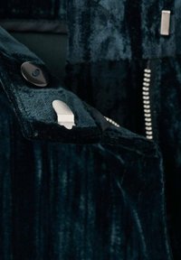 Lähikuva tummasta samettitakkihihasta, jossa on suuri musta nappi ja hopeinen kiinnitys, vetoketju näkyy taustalla.