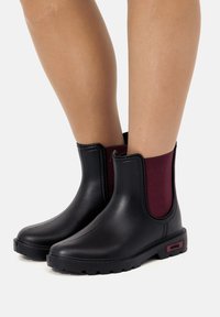 Stivali impermeabili neri alla caviglia con pannello elastico bordeaux, suola in gomma texturizzata e finitura liscia. Presentano hardware minimale e un branding discreto.