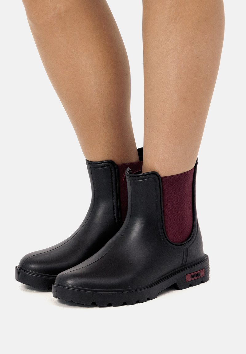 Stivali impermeabili neri alla caviglia con pannello elastico bordeaux, suola in gomma texturizzata e finitura liscia. Presentano hardware minimale e un branding discreto.