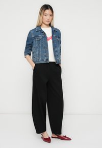 Kvinne står med hendene i lommene, iført en blå denimjakke, hvit skjorte med røde lepper trykk, svarte vide bukser, og røde spisse sko.