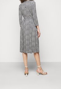 Kleid mit floralem Muster in Schwarz und Weiß mit langen Ärmeln. Das Design hat eine taillierte Taille und einen ausgestellten Rock, kombiniert mit beigen, schnürbaren Absatzschuhen.